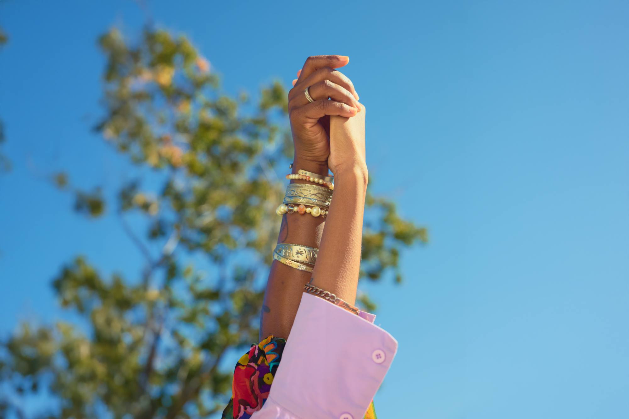 Two hands clasped together against a clear blue sky, adorned with an assortment of silver and beaded bracelets. A hint of a colorful floral garment adds vibrancy, while blurred tree branches create a natural background.