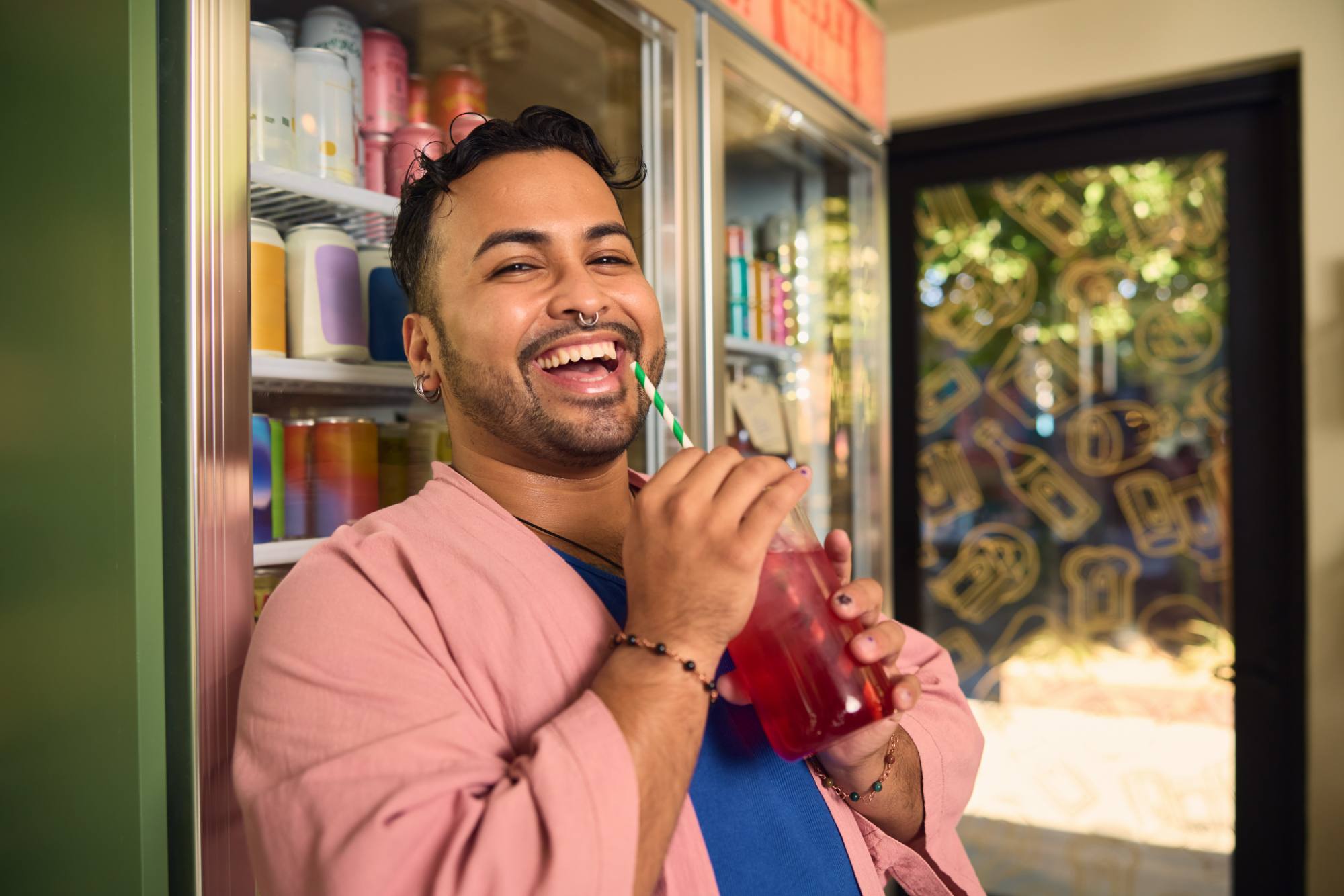 A cheerful person wearing a pink shirt enjoys a refreshing red drink with a striped straw, standing next to a refrigerator stocked with colorful beverages. A decorated glass door in the background adds a vibrant, playful touch to the scene.