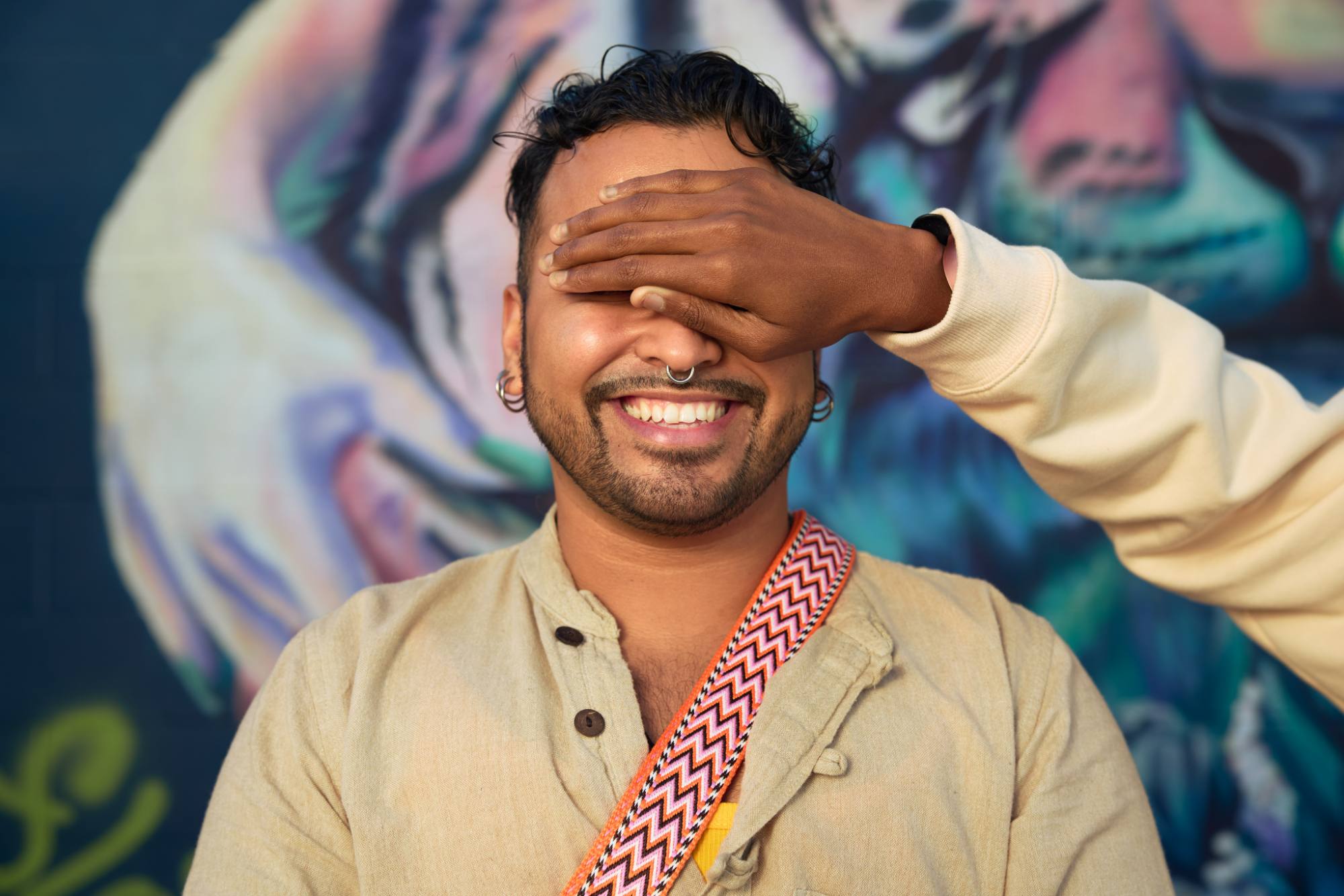 A smiling person with a nose ring and earrings stands in front of a vibrant mural, wearing a beige shirt and a colorful patterned strap across their shoulder. A hand playfully covers their eyes, adding a sense of fun and surprise to the scene. The mural’s abstract design creates a lively and artistic backdrop.