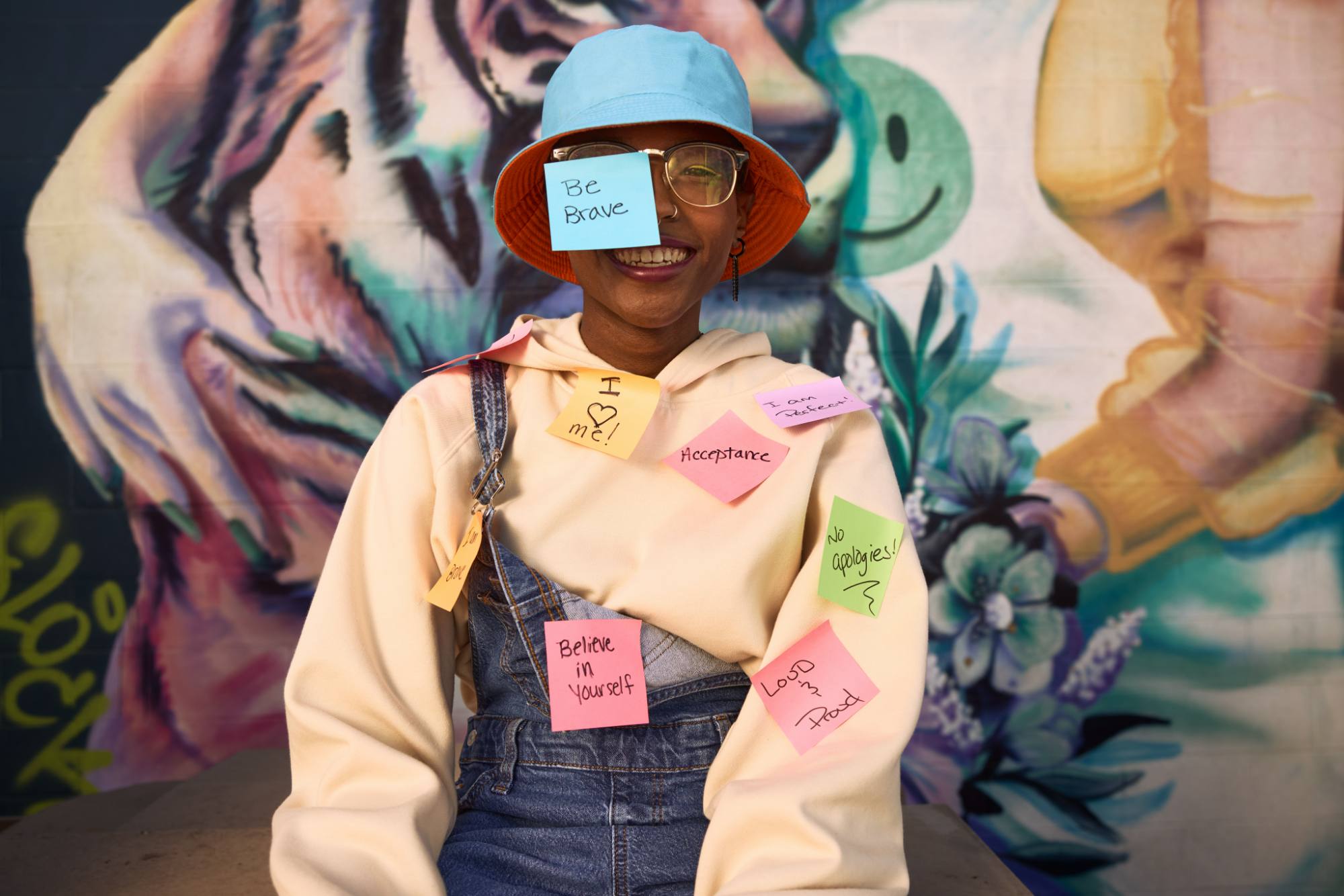 A person wearing overalls, a hoodie, and a bucket hat smiles joyfully with colorful sticky notes attached to their outfit and face. The notes display empowering messages like "Be Brave," "Acceptance," and "Believe in Yourself." A vibrant mural featuring abstract designs and flowers is visible in the background, enhancing the uplifting and creative atmosphere.