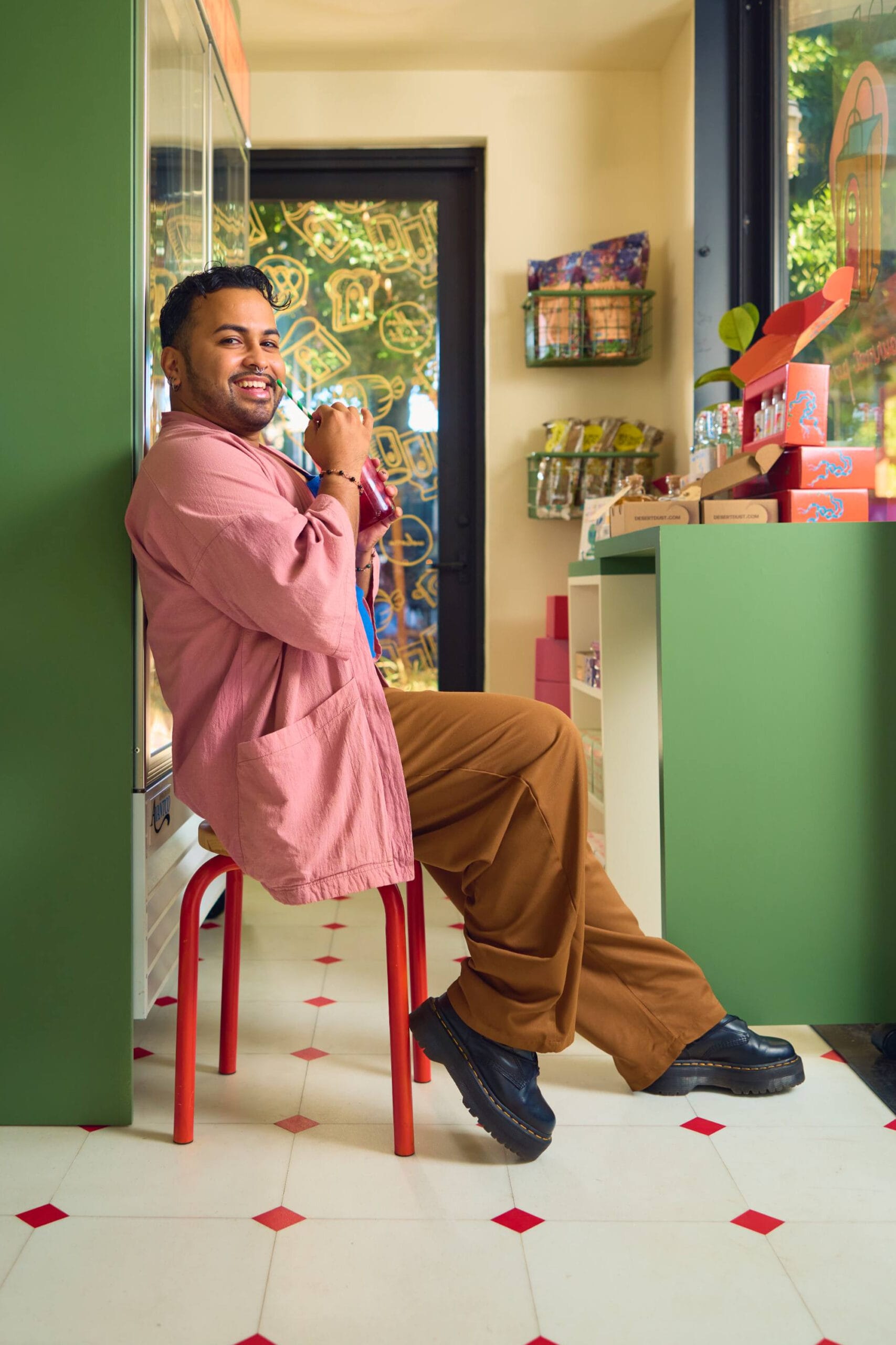 A cheerful person sitting on a red stool in a cozy shop, holding a drink and smiling at the camera. They are wearing a pink shirt and brown pants, with a display of snacks and colorful packaging in the background. The scene is warmly lit, with a playful pattern visible on the shop's glass door.