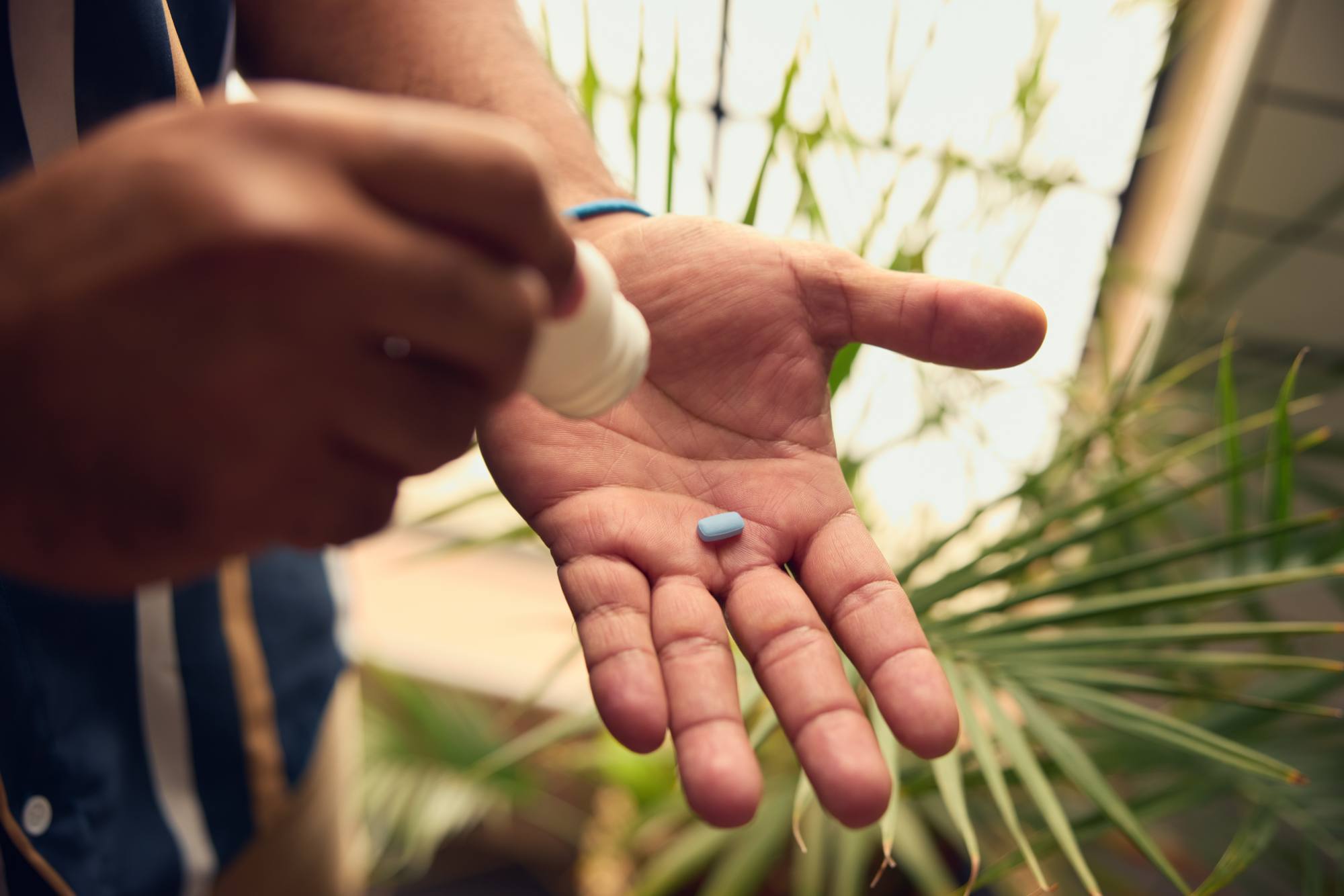 A close-up of a person pouring a single blue pill from a bottle into their palm, set against a background of green plants and natural light. The scene conveys a moment of self-care or medication, with a focus on health and wellness in a serene outdoor setting.