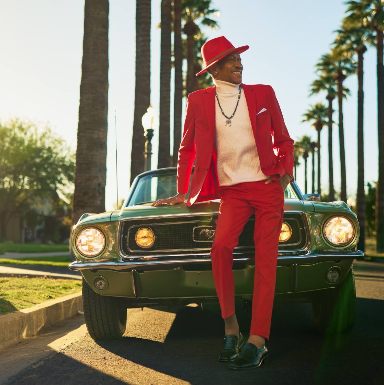 A stylish person dressed in a bold red suit, matching hat, and white turtleneck leans confidently on a classic green Ford Mustang. The backdrop features a sunny street lined with tall palm trees, creating a retro and vibrant vibe. The scene exudes charisma, individuality, and timeless cool.