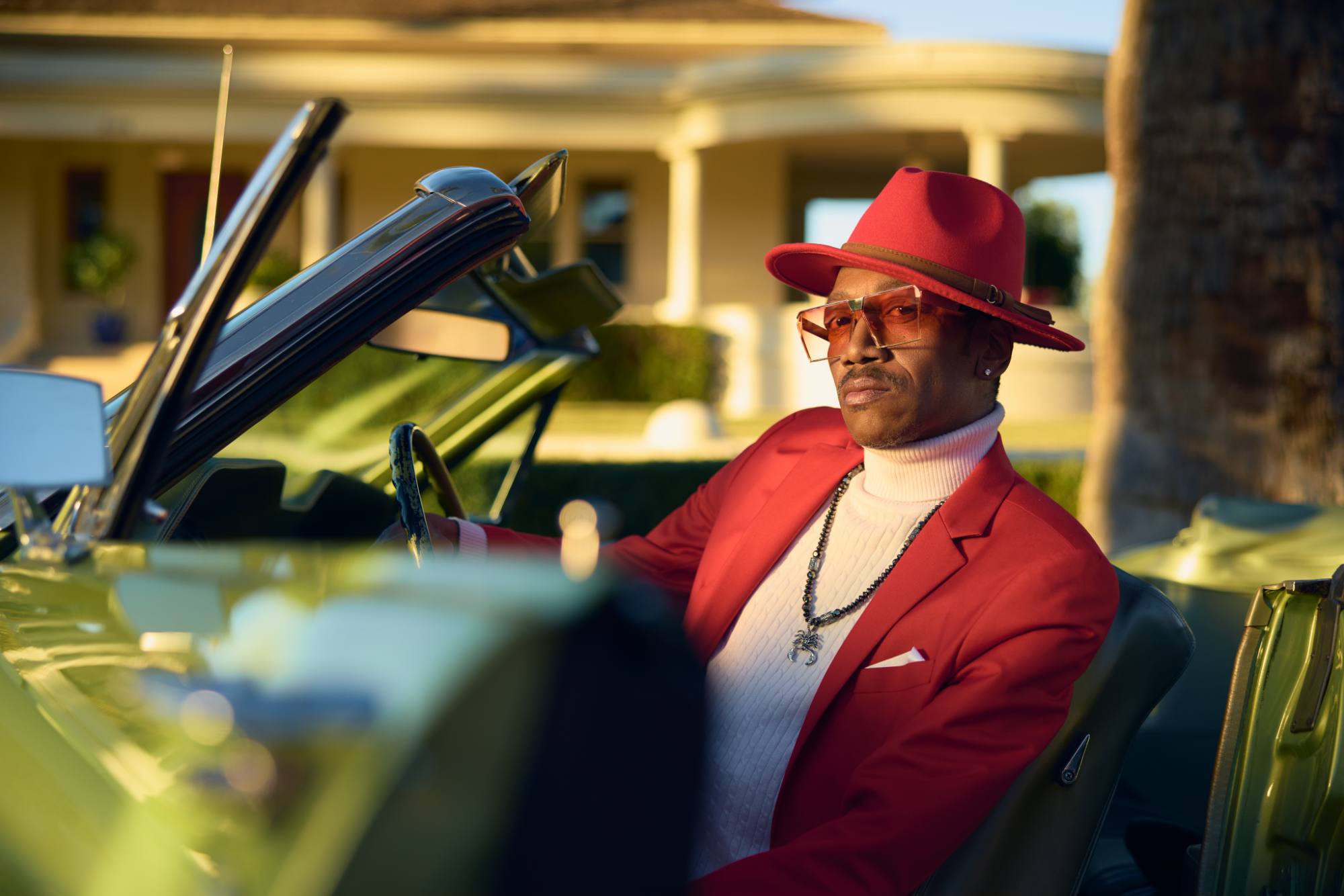 A stylish man sits confidently in a vintage green convertible, wearing a red suit jacket, white turtleneck, matching red hat, and tinted sunglasses. The sunlight casts a warm glow, highlighting the luxurious and sophisticated atmosphere, with a suburban house blurred in the background.