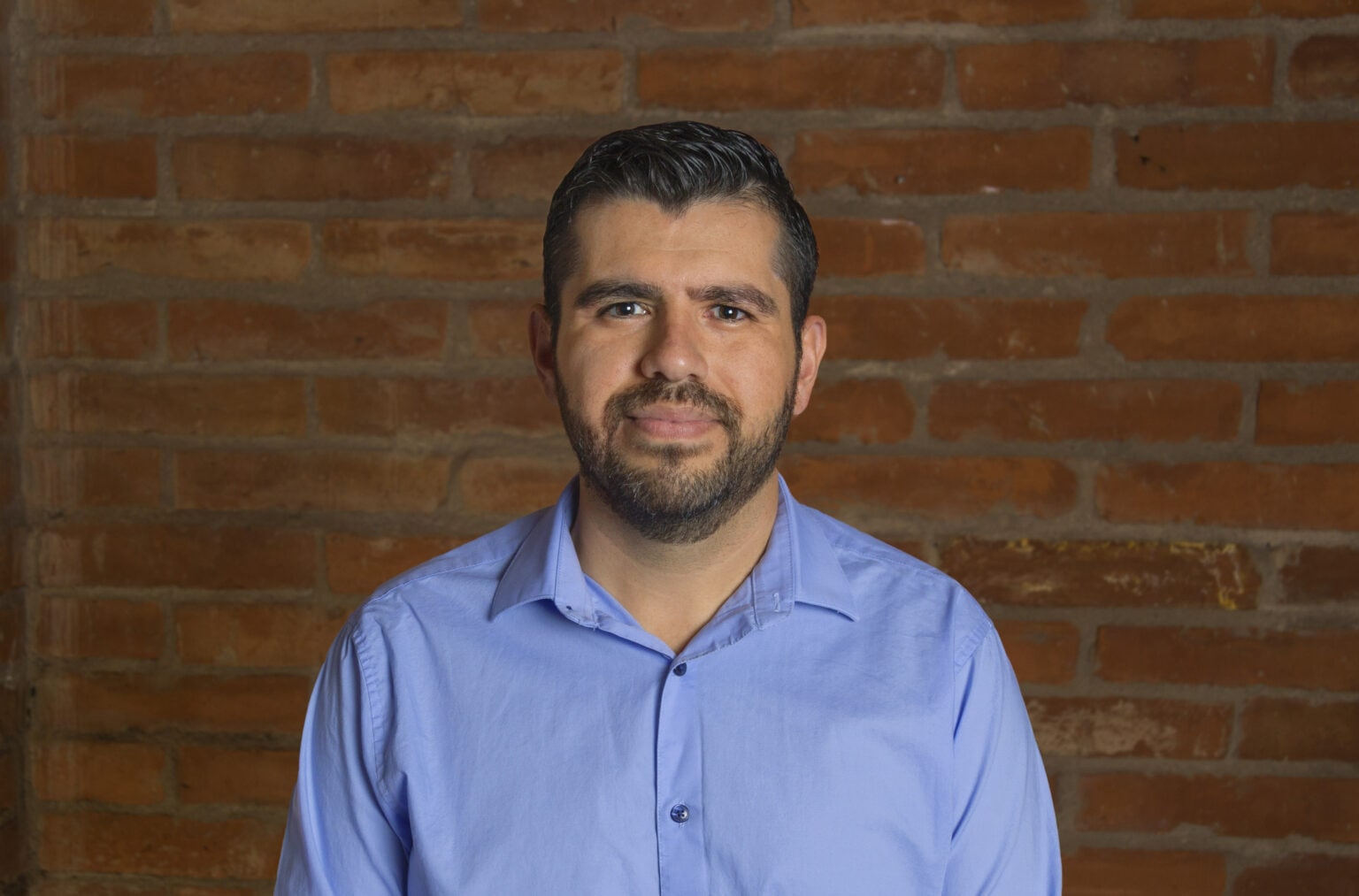 Dr Jose Rodriguez Garcia, A portrait of a man with neatly combed dark hair and a beard, wearing a light blue collared shirt. He stands against a rustic red brick wall, with warm lighting highlighting his confident and approachable expression.