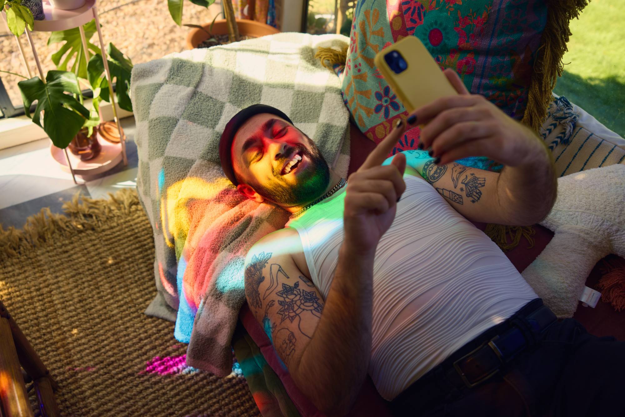 A bearded individual reclines on a cozy setup with patterned blankets and pillows, smiling while using a smartphone. Sunlight filters through the window, casting colorful rainbow patterns across their face and body, creating a warm and vibrant atmosphere surrounded by indoor plants and bohemian decor.