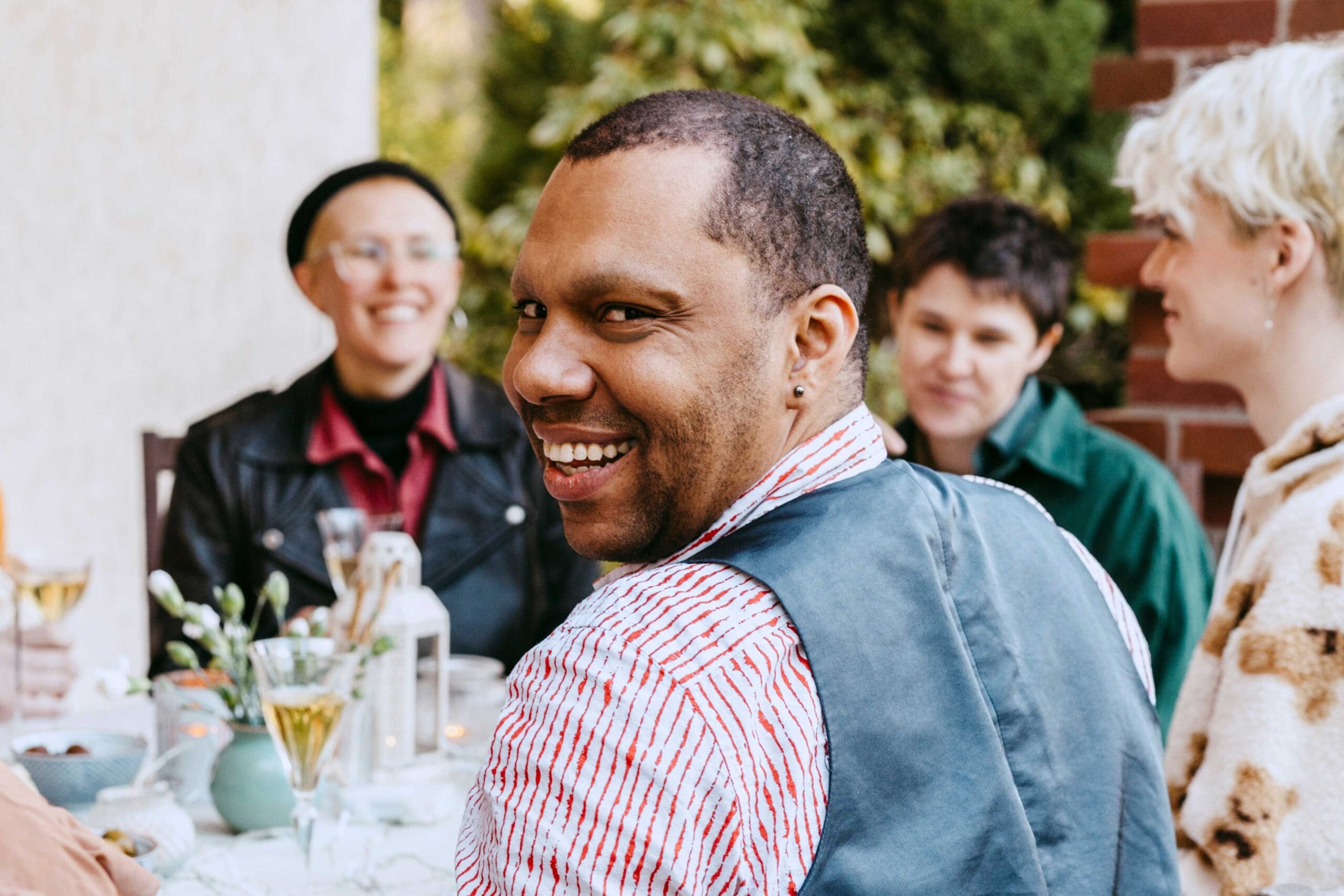 A joyful outdoor gathering featuring a smiling man in the foreground wearing a striped shirt and vest, turning to look at the camera. In the background, three other people are seated around a table decorated with flowers, candles, and glasses, enjoying a relaxed and friendly atmosphere.