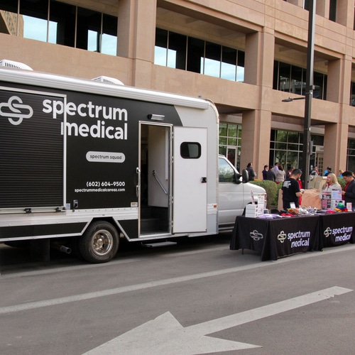 A mobile medical van labeled "Spectrum Medical" is parked on a street, with an open door providing a view of its interior. Nearby, tables set up with "Spectrum Medical" branding display informational materials and small items, while several people interact in the background. The scene highlights a community health outreach or event in an urban setting.