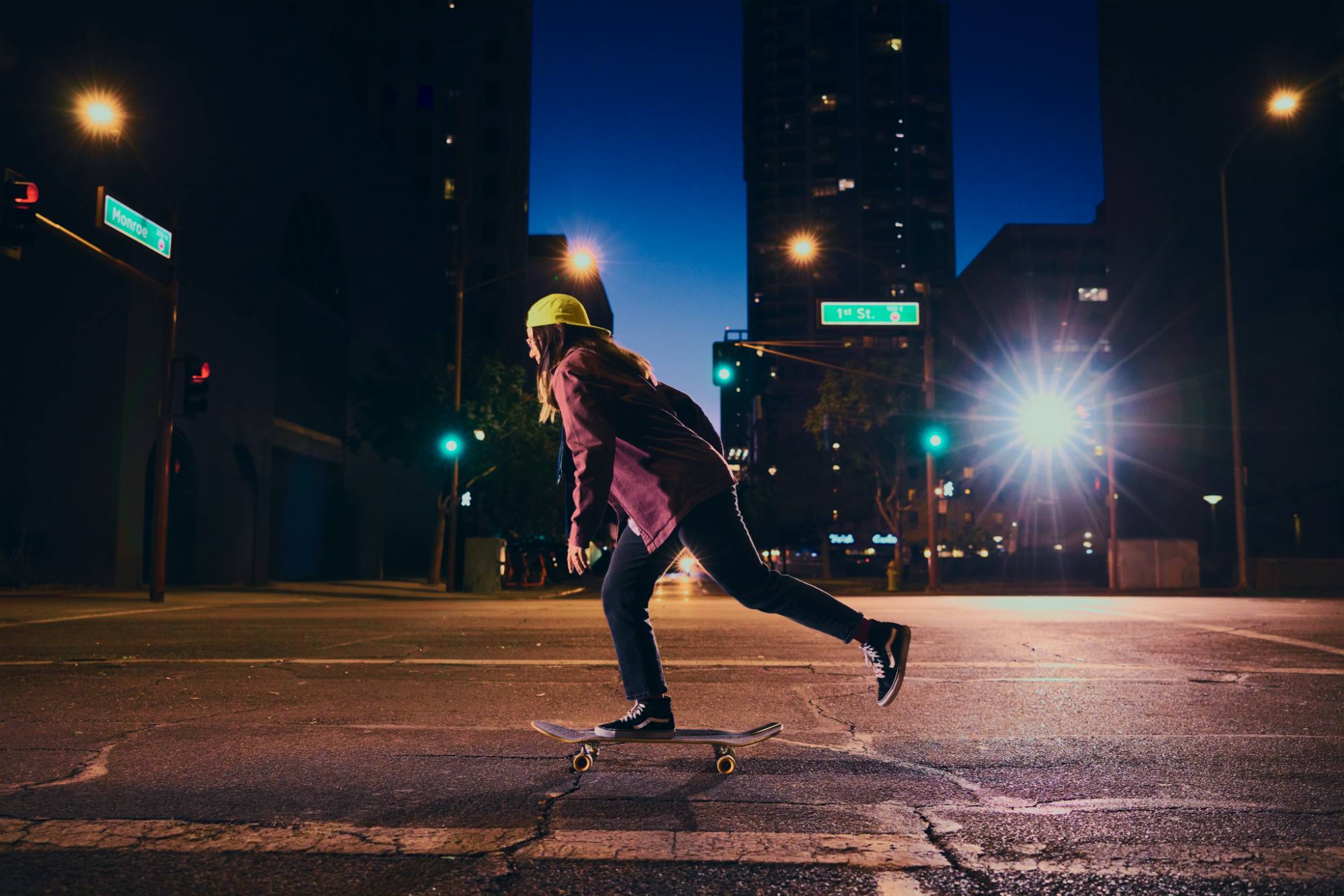 A skateboarder wearing a yellow cap and casual streetwear glides through an empty urban intersection at night. The city lights and glowing street signs, including "Monroe" and "1st St," illuminate the scene against a deep blue sky. The image captures a sense of freedom, adventure, and urban energy.