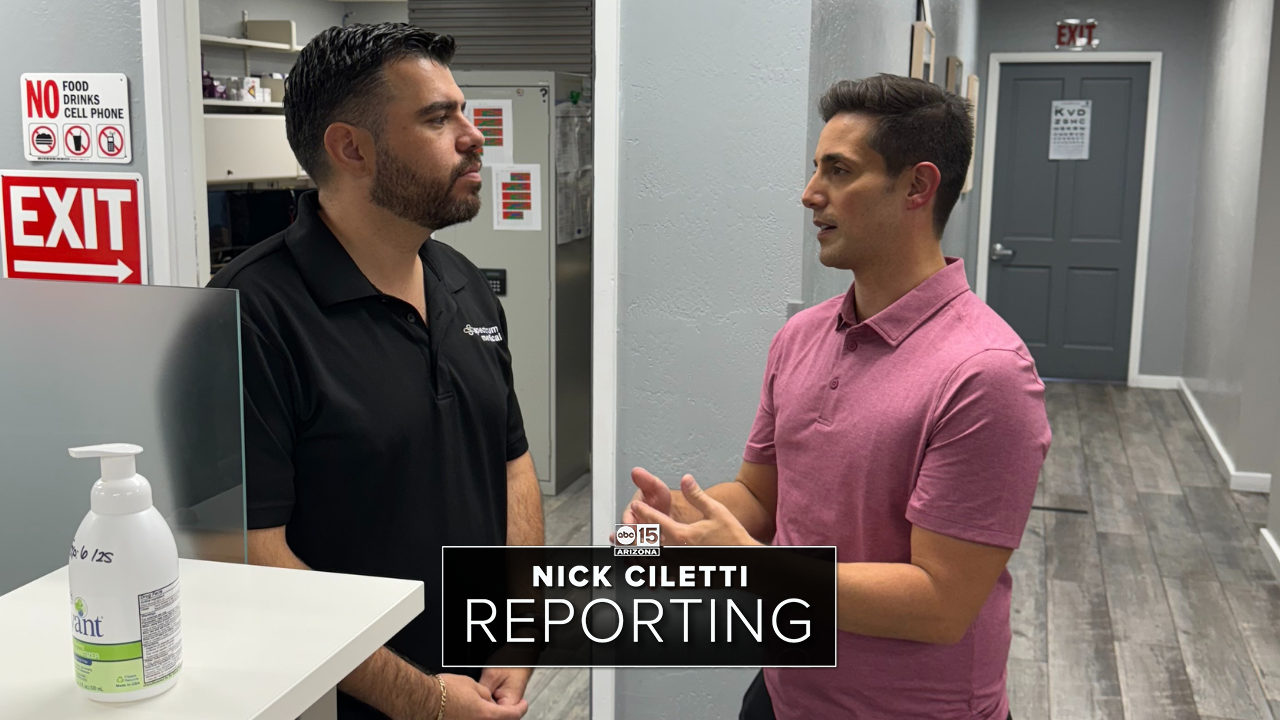 Two men are engaged in conversation in a clinical office setting. One, wearing a black polo with "Spectrum Medical" branding, listens attentively, while the other, dressed in a pink polo, gestures as he speaks. The text overlay reads "Nick Ciletti Reporting," indicating a news segment or interview for ABC15 Arizona.
