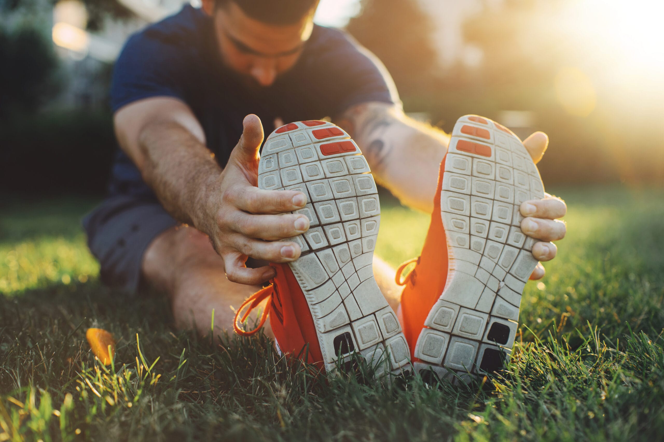 A person sits on grass in a sunlit outdoor setting, stretching forward to touch their toes while wearing bright orange athletic shoes. The focus is on the soles of the shoes, which show a textured grip, with a warm, golden glow in the background emphasizing the serene and active atmosphere.