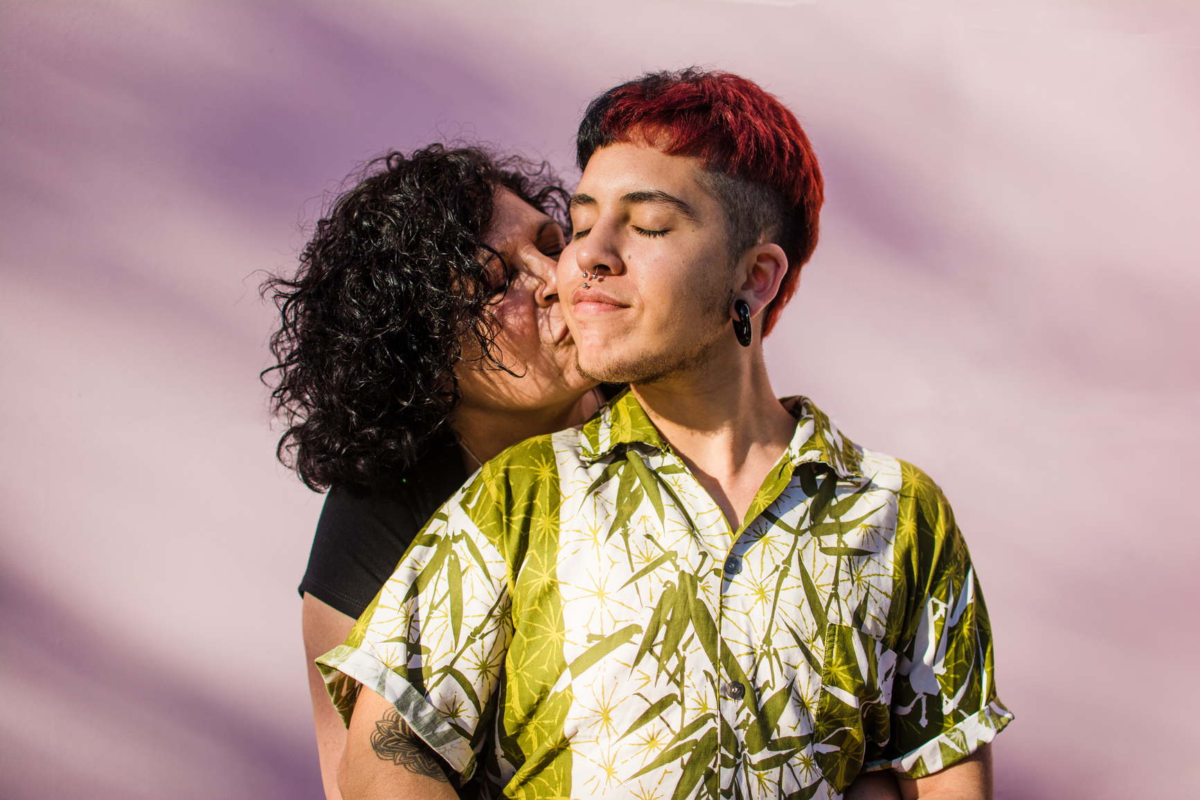 Young latino transgender man posing against a pink wall and holding a pinwheel with the trans rights flag colors. His mother is hugging him lovingly.