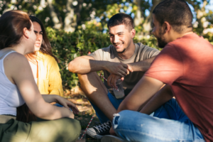 A group of happy people sit together and converse while cross-legged on the ground outside at a park.