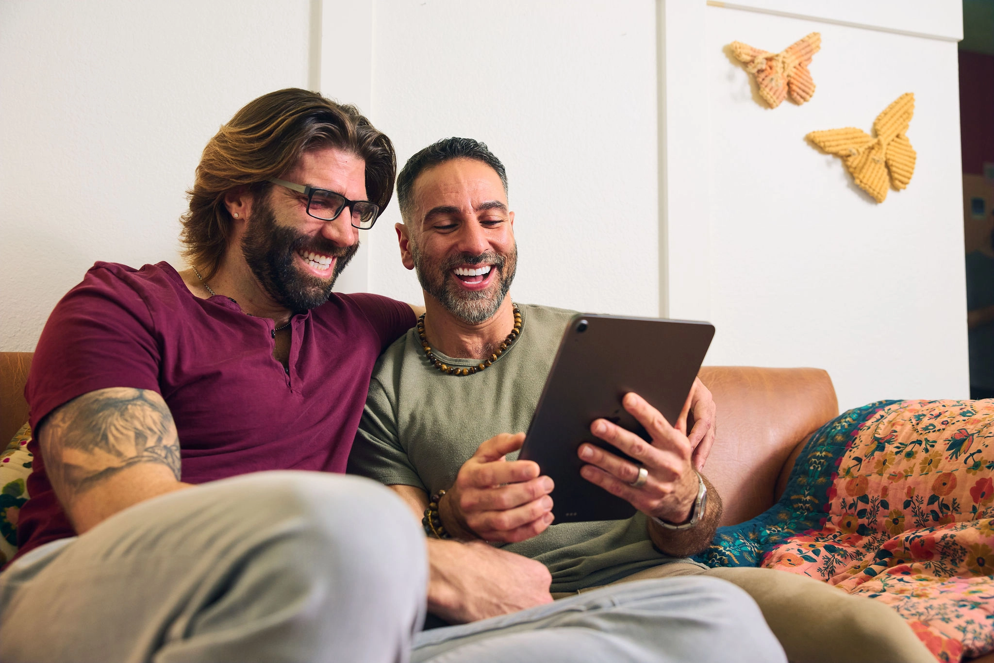 A gay couple leans into each other on a couch while smiling at the display on an iPad that we cannot see.