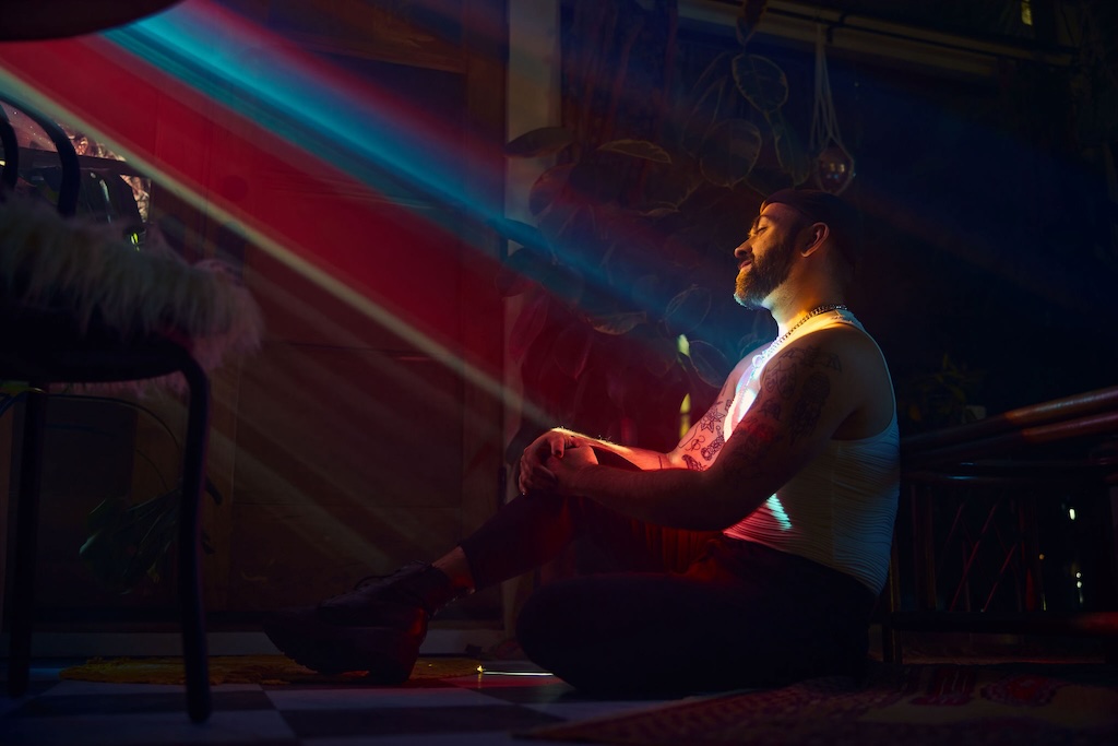 A man sits in a dark room, illuminated by a beam of rainbow light.