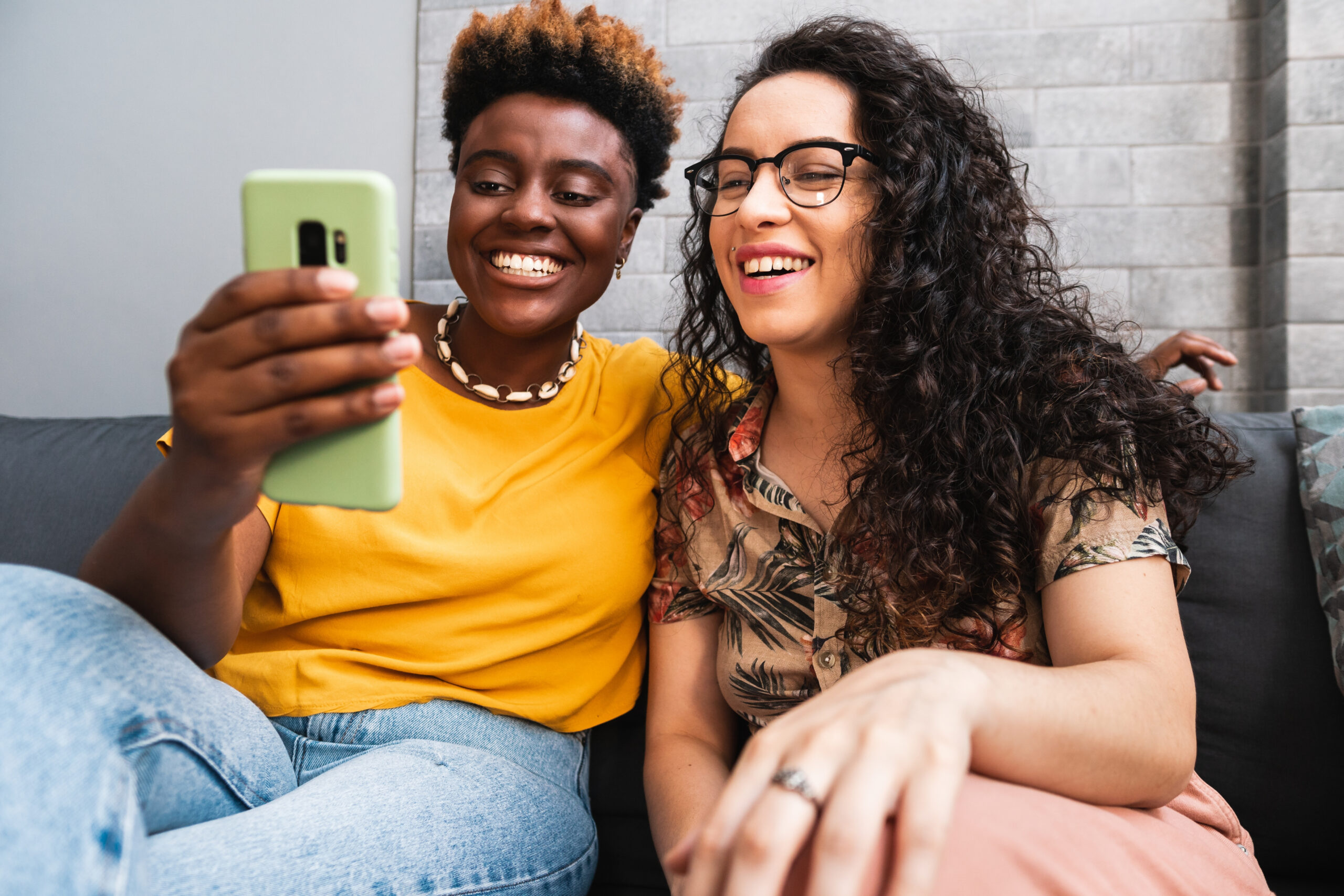 Two smiling women sitting on a couch together viewing a phone. We can't see what's on the phone.