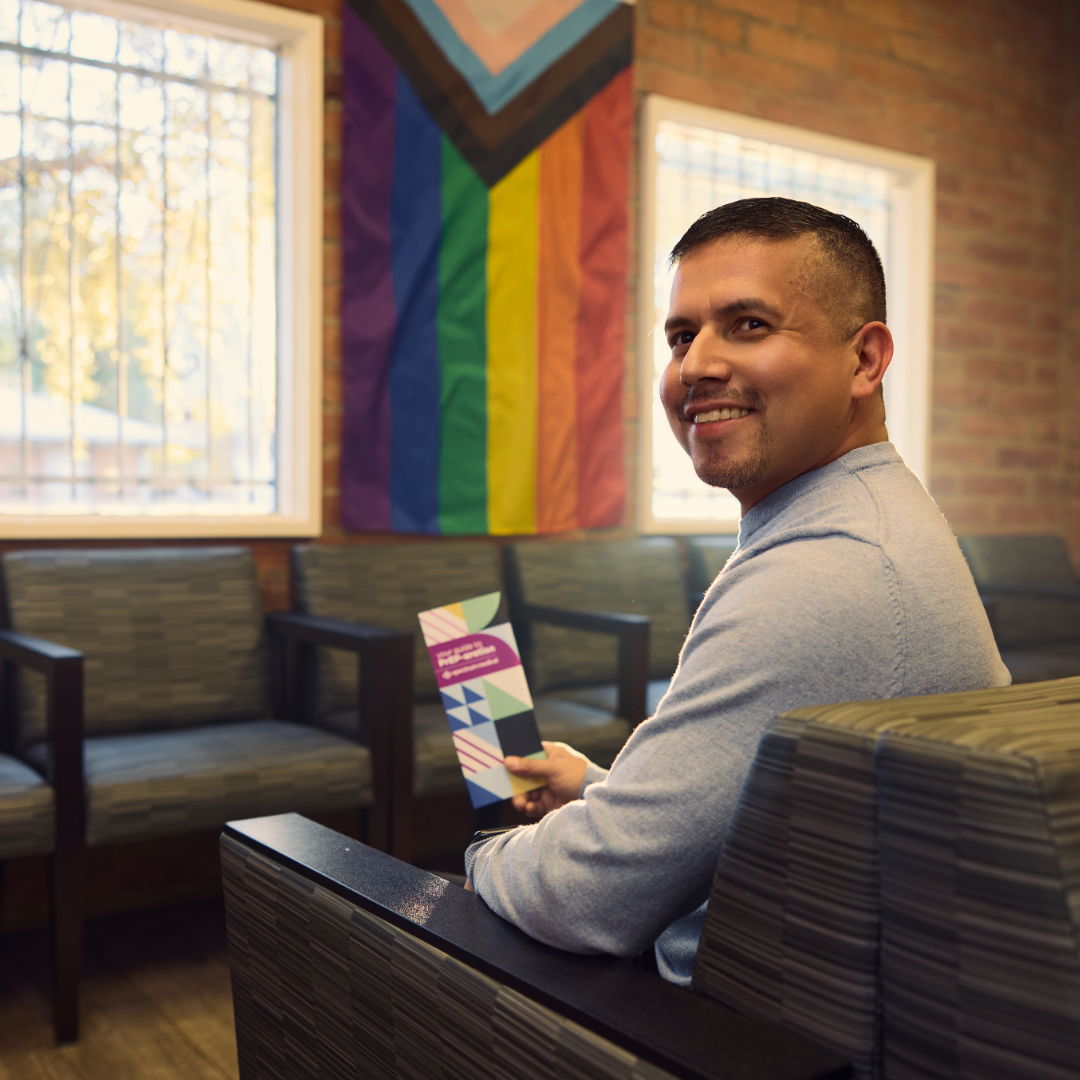 A person sits in a clinic waiting area, smiling and holding a brochure, with a rainbow Pride flag displayed on the wall behind them, creating a welcoming and inclusive environment.