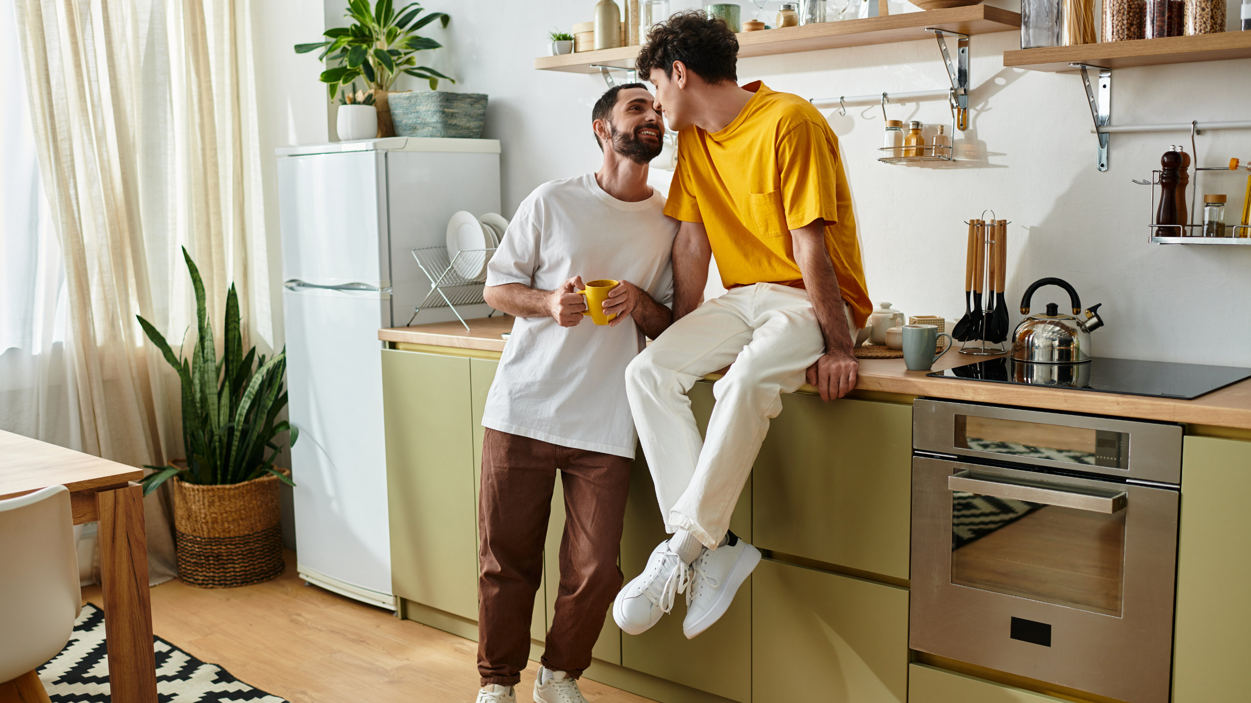 Two men share a kiss in a bright modern kitchen; one sits on the counter in a yellow shirt, the other stands in white.