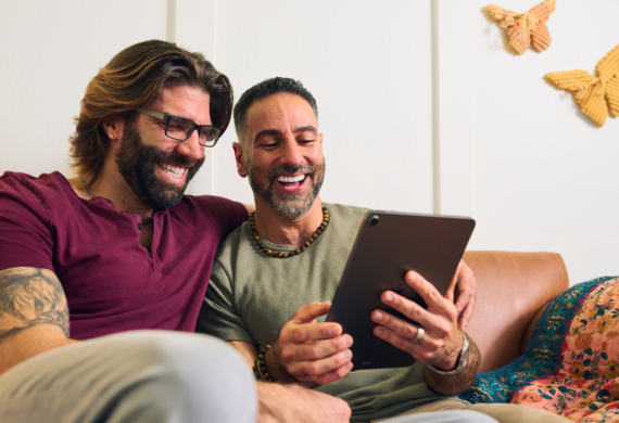 Two men sit together on a couch, smiling and looking at a tablet, sharing a relaxed and happy moment.