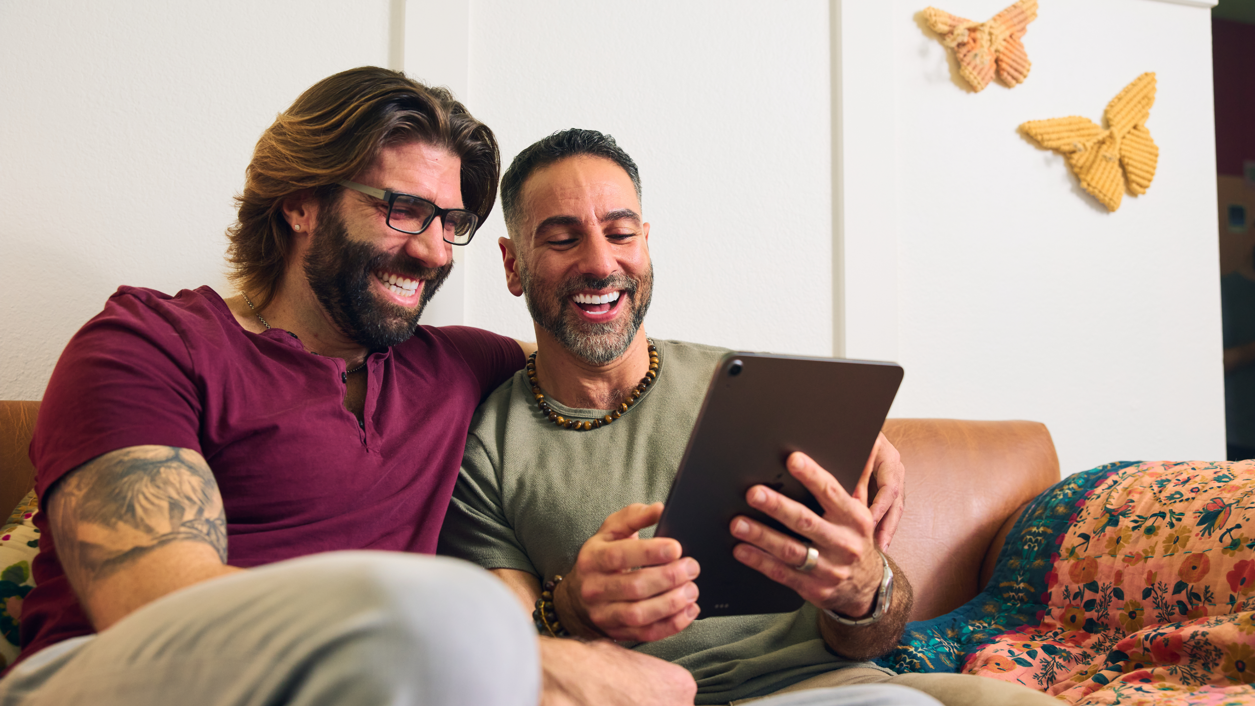 Two men sit together on a couch, smiling and looking at a tablet, sharing a relaxed and happy moment.