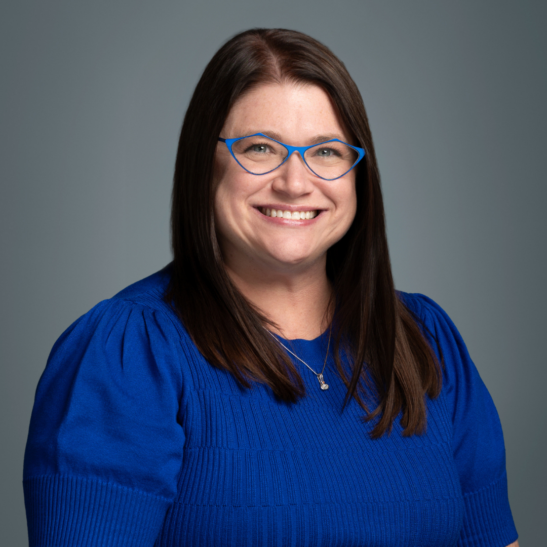 Professional headshot of a smiling woman with dark hair and blue glasses wearing a royal blue top