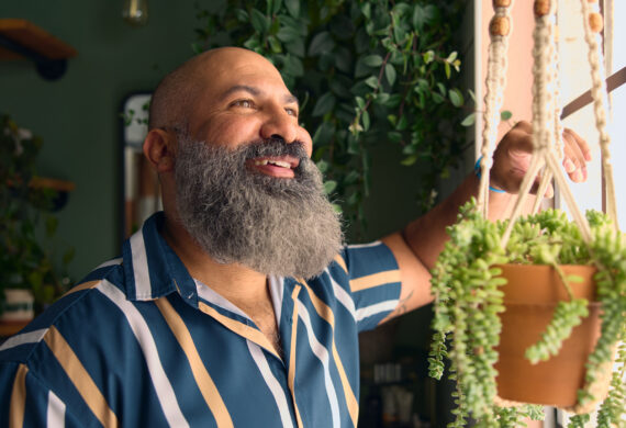 A smiling man with a long gray beard stands indoors beside a window, looking outside while holding a hanging succulent plant.