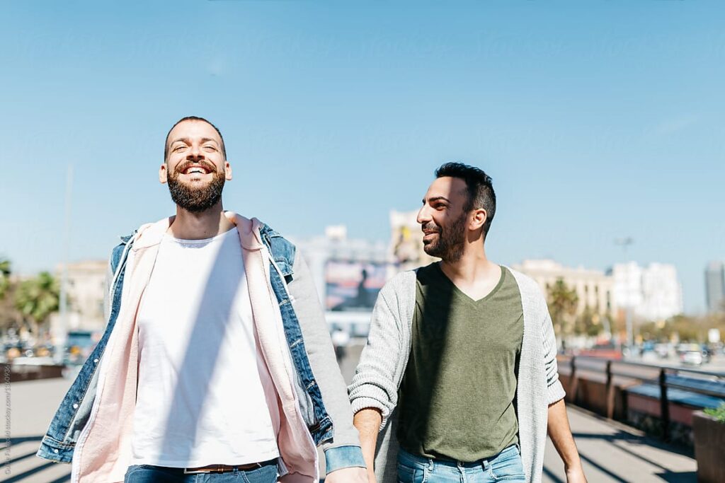 Two men walk arm in arm along a sunny seaside promenade, smiling at each other.