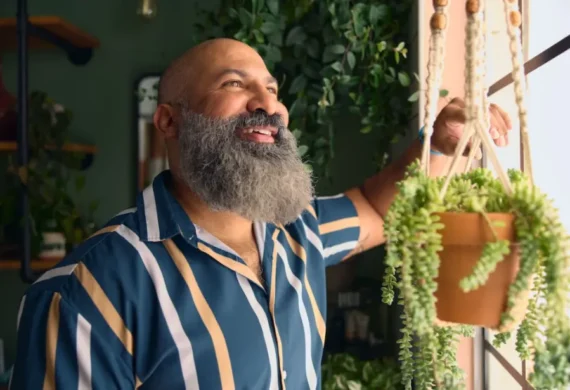 Bearded man in a blue and tan striped shirt smiles while adjusting a hanging potted plant indoors in a greenery shop.