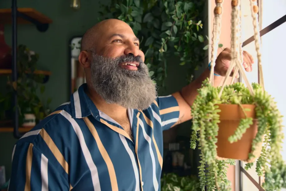 Bearded man in a blue and tan striped shirt smiles while adjusting a hanging potted plant indoors in a greenery shop.