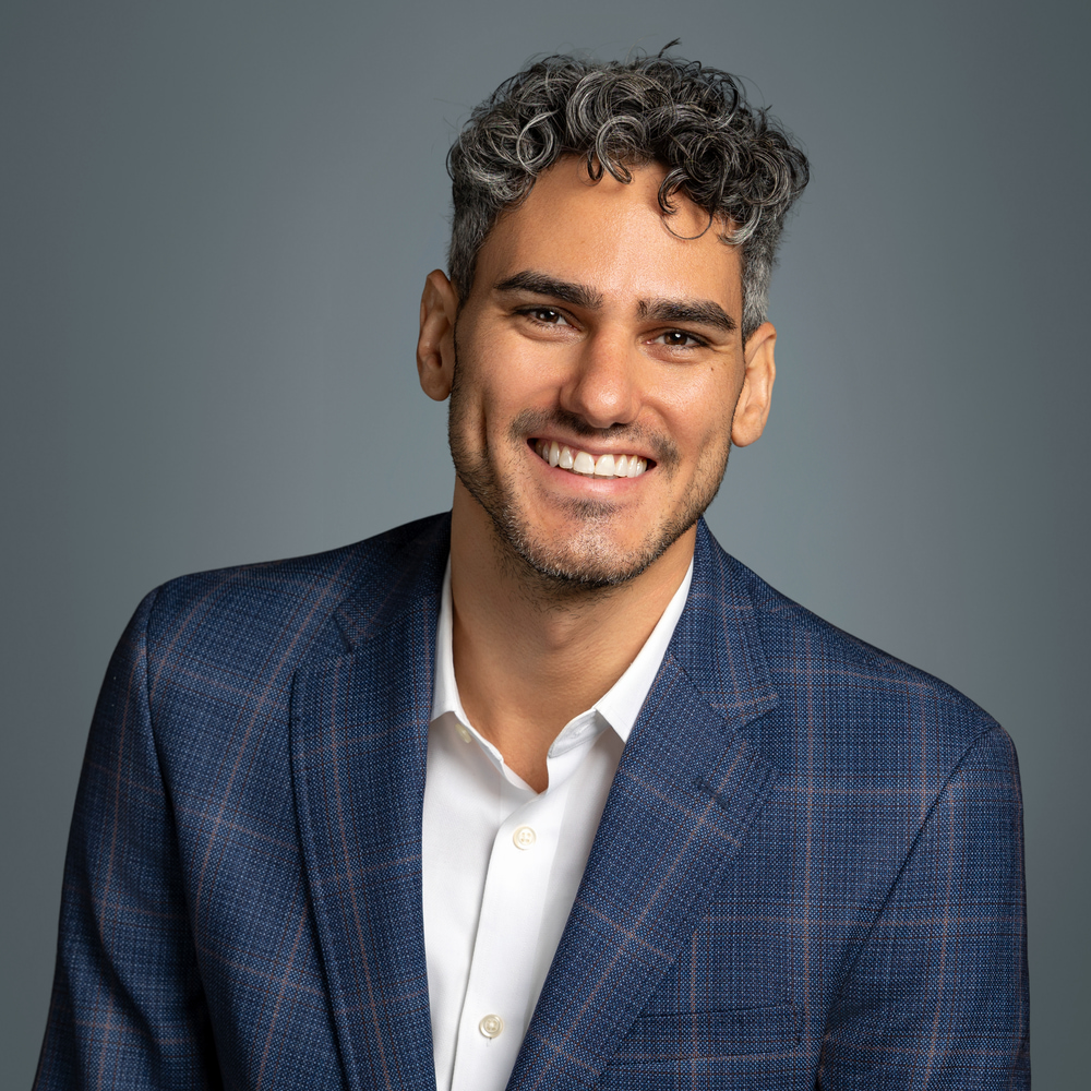 Portrait of a smiling man in a navy check blazer and white shirt, against a gray studio backdrop.