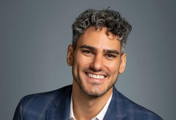 Close-up portrait of a smiling man with gray-curled hair wearing a blue plaid blazer against a gray background, cropped at the shoulders.