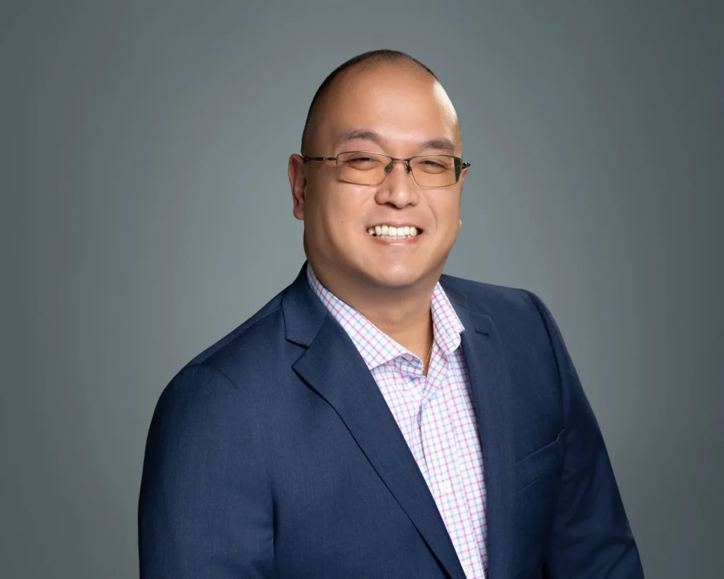 Professional headshot of Van Lieu. He is smiling in a navy suit jacket and checkered shirt, wearing glasses, against a gray background.